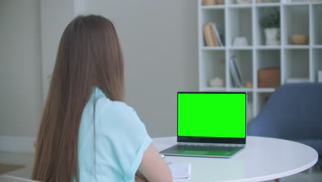 Woman-sits-at-desk-in-bedroom-she-looks-at-laptop-green-screen-and-talks-to-someone-over-internet-video-communications-sometimes-taking-notes-in-notebook.-Close-up